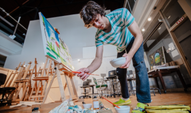 Mason Terra holds a brush and bowl as he paints on a canvas 