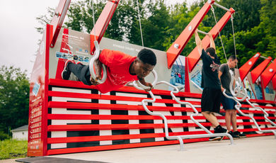 A man wearing a red shirt hangs on rings on outside fitness court 