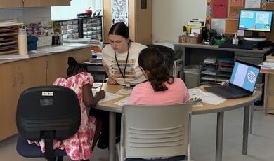 A student works with children while all are sitting at a table in a classroom.
