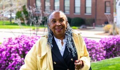 Sheryl Royster stands in front of purple flowers while wearing a yellow jacket.