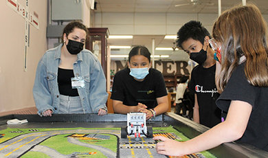 A BSU student helps fifth graders test a robot on a mat.