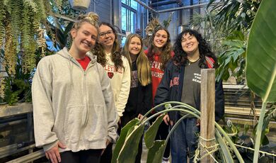 Women stand in a greenhouse next to plants 