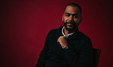 portrait photo of Harold Tavares sitting in a chair in front of a red backdrop