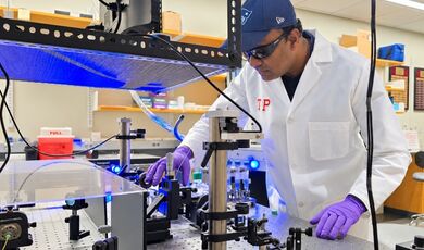 A professor in a white lab coat works at a table filled with optical equipment.