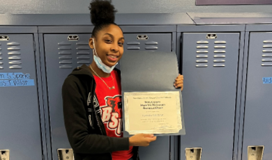 Tyeisha Gilchrist stands in front of lockers with award saying she is first Martin Richard Foundation scholar