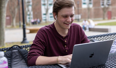 A man wearing a red shirt sits looking at his computer screen and smiles