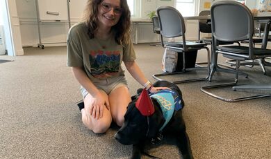 A woman wearing glasses sits down next to a dog wearing a party hat 