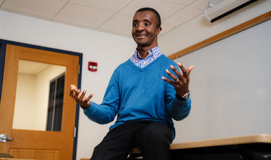 Wayne Magee is sitting on a desk wearing a blue sweater smiling with his hands held up in the air 