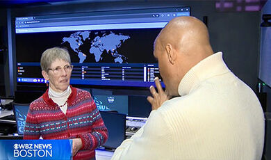 A journalist interviews a professor in the cyber range with a large digital display in the background showing a map of the world.