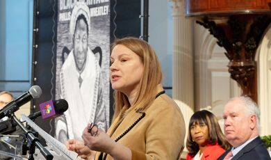 Emily Field speaks from behind a podium with a picture of the Phillis Wheatley stamp behind her.