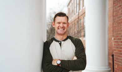 A man smiles with hands folded in front of chest leaning on a pillar 