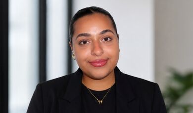 Headshot of Zainab standing in front of windows and a wall.