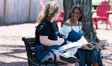 Two smiling students sitting on a bench outside, with a laptop and notebook