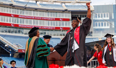 A BSU College of Business graduate crossing the stage at Commencement smiles and raises his arm while shaking hands with Dean Jeanean Davis-Street