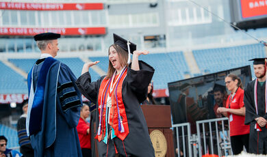 A female BSU graduate crossing the stage at Commencement smiling and raising the roof with her arms