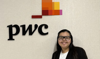 Glendys Bonilla, '27, standing next to a wall with the Price Waterhouse Cooper logo on it. She has long straight dark hair and is wearing black rim glasses and a black blazer over a white top