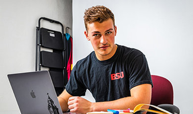 Ryan Kuczer, '23, sitting at an open laptop wearing a black BSU t-shirt