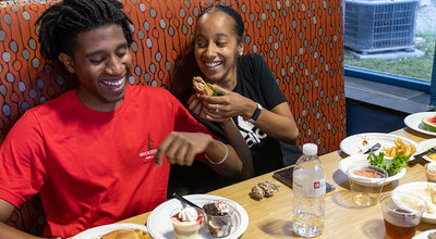 Two students with plates of food and pudding cups laughing in a booth at The Bear's Den