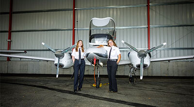 Two students stand next to an airplane inside a hangar.