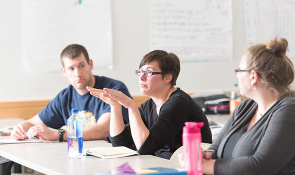 3 BSU graduate students sitting at a table in an education classroom, one is speaking while the other 2 listen