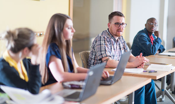 4 students sitting at tables in a classroom, 2 with laptops open, the other 2 with notebook and papers in front of them, one of the male students speaking