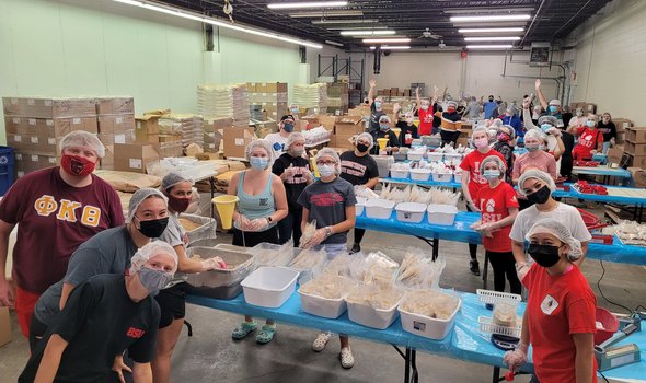Photo of BSU student volunteers at End Hunger New England packing meals for folks experiencing homelessness. They are spaced out across 4 tables in groups, and are wearing masks, gloves and hairnets and looking at the camera