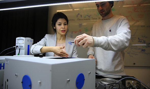 Dr. Elif Demirbas working with a student in front of a large piece of equipment. The student is holding a small piece of blue plastic with a pair of tweezers
