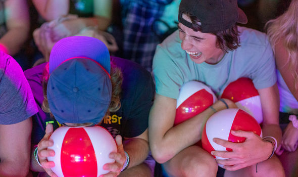 2 BSU first year students sitting next to each other at Crimson Days event. One, wearing a blue baseball cap, has his forehead resting on top of a small red and white inflatable ball while the second student with a black baseball cap worn backwards holds 3 small inflatable balls while looking at the first and laughing