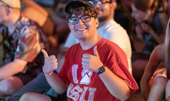 A BSU student smiling and giving thumbs up, wearing glasses, a baseball cap and an I paw BSU tshirt, sitting on a floor surrounded by other students