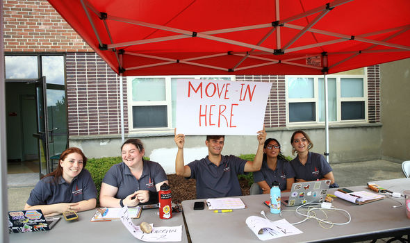 5 BSU students sit smiling at tables under a red canopy in front of a dorm with one holding a sign that says MOVE-IN HERE