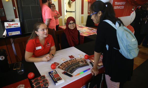 2 students sit behind a table with materials on it talking to another student leaning over the table