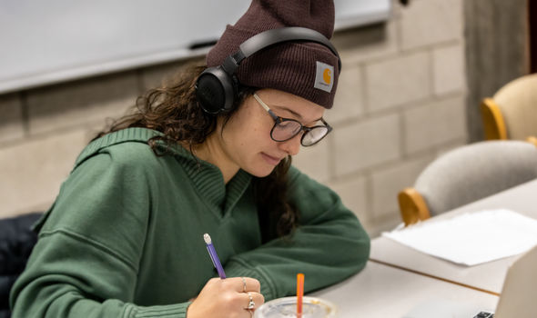 a female student with long brown hair wearing glasses, a brown winter hat, headphones and green hoodie sits at a table and writes on paper while a laptop sits open in front of her