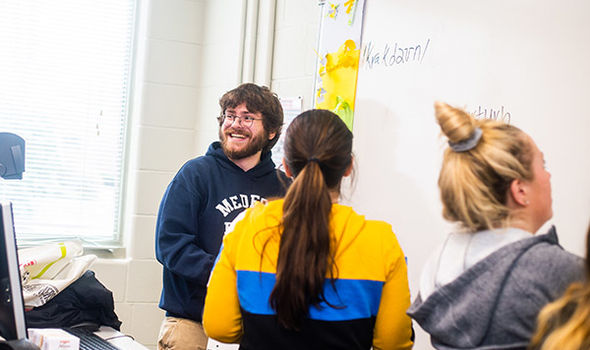 students in phonetics class working at the white board while one looks toward the classroom smiling