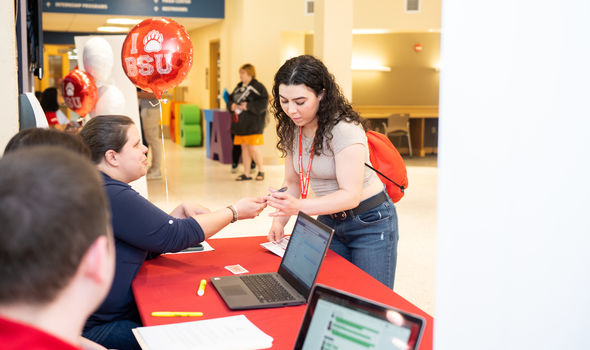 BSU student showing her phone to a staff member sitting at a table with a red cloth, balloon and laptops on it