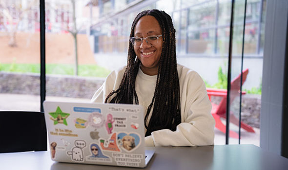 A student smiling and working on a laptop in bright, windowed space