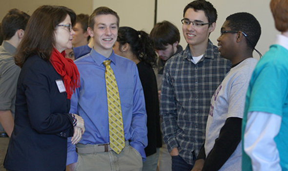 3 high school students talk to a teacher with other students in the background