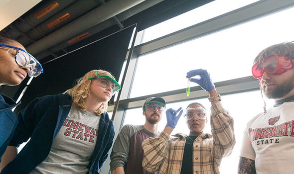 A student holds up a test tube of green liquid; a group of students and their professor look on