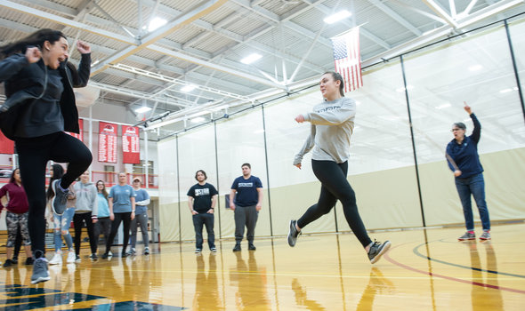 physical education class in the gym showing 2 students racing while others watch and professor referees