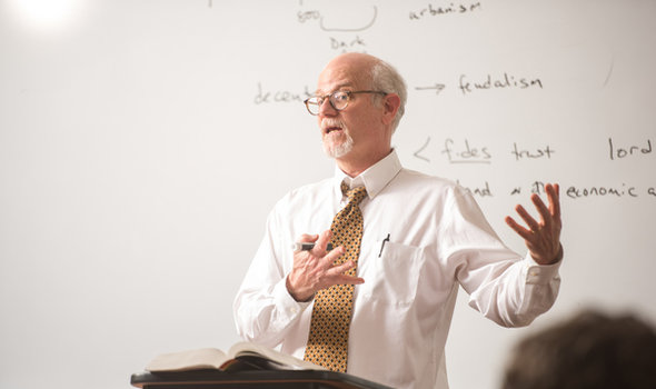 History professor gesturing while talking to his class in front of a white board with notes on feudalism