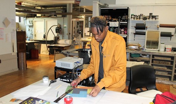 a BSU student standing at a large table in a studio of the Art building measuring out and cutting blocks of color on a paper