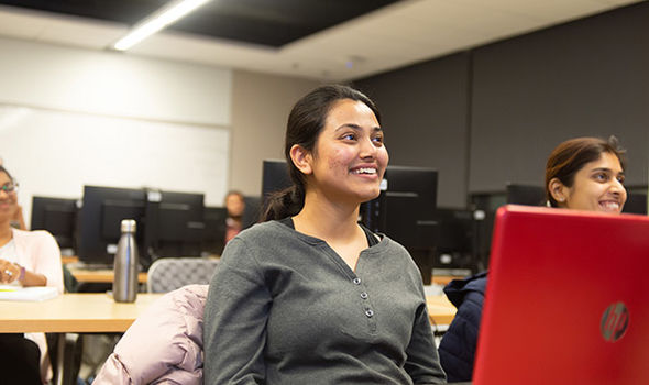students sitting at tables in classroom with their laptops open smiling and looking at front of class
