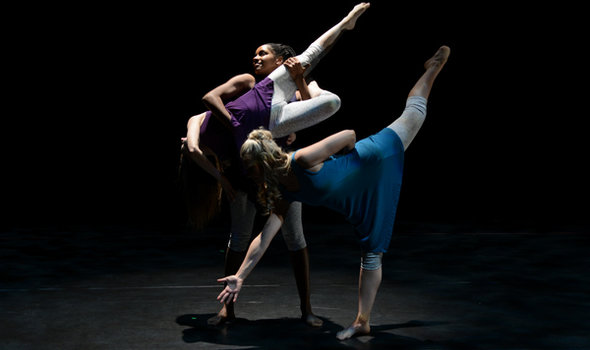 3 female dancers spotlighted on a dark stage all wearing light gray cropped leggings and different colored tunic tops; 1 standing holding another whose head dips towards the floor, her left leg pointed up and the other bent in while the third dancer stands in front of them with her right arm pointed toward the ground, left arm behind her, left leg pointed up and extended, standing on her straight right leg