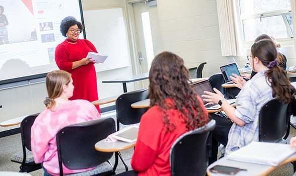 Professor Sheena Manuel smiling and holding a notebook in front of a projector screen at the front of the classroom while she listens to a student speaking in class