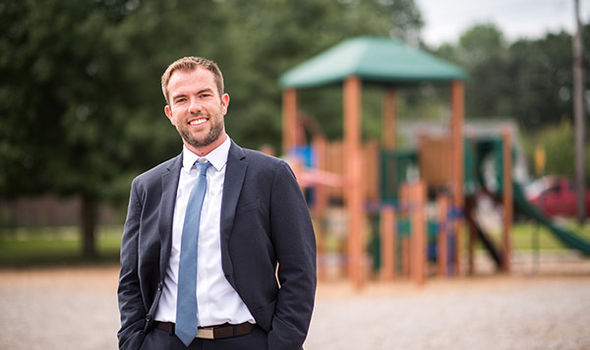 BSU Ed Leadership grad and Elementary School Principal Edward Paris standing on a playground wearing a dark gray suit, white button down shirt and light blue tie