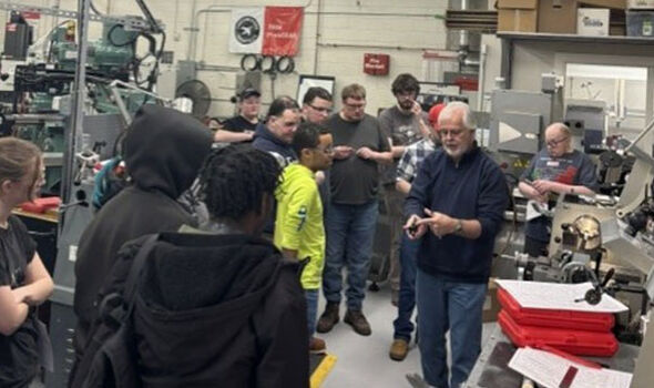 In a machine shop an older man shows something to a group of students standing around him