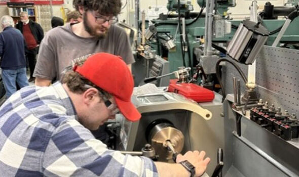 In a machine shop a young man operates a machine while another looks on