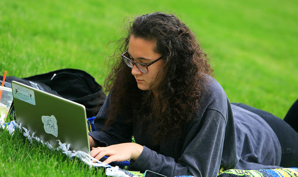 A student working on a laptop while laying on the grass