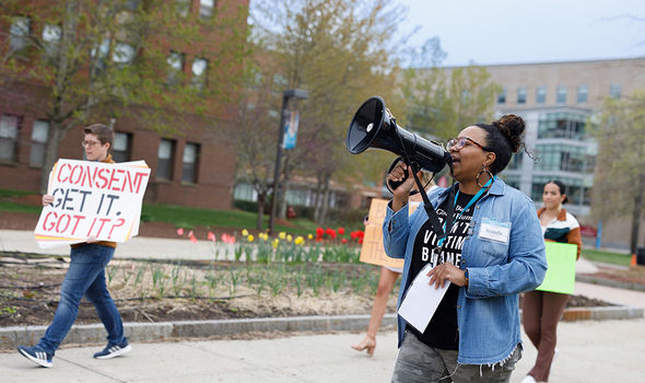 A student organizer speaks through a megaphone as students march in a Take Back the Night rally; in the background, a person holds a sign that says, "Consent: Get it. Got It?"