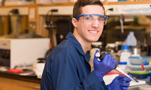 BSU Chemistry major sitting and smiling in a lab wearing safety glasses, a dark blue lab coat and gloves