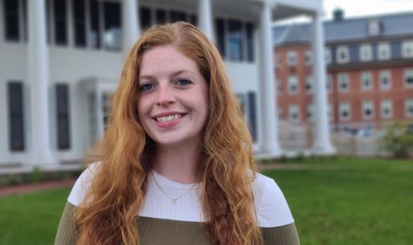 Sarah Murphy smiles at the camera in front of 161 summer street. She has red, wavy hair which she is wearing down and is wearing a cream colored swaeter with an olive green stripe across the chest.